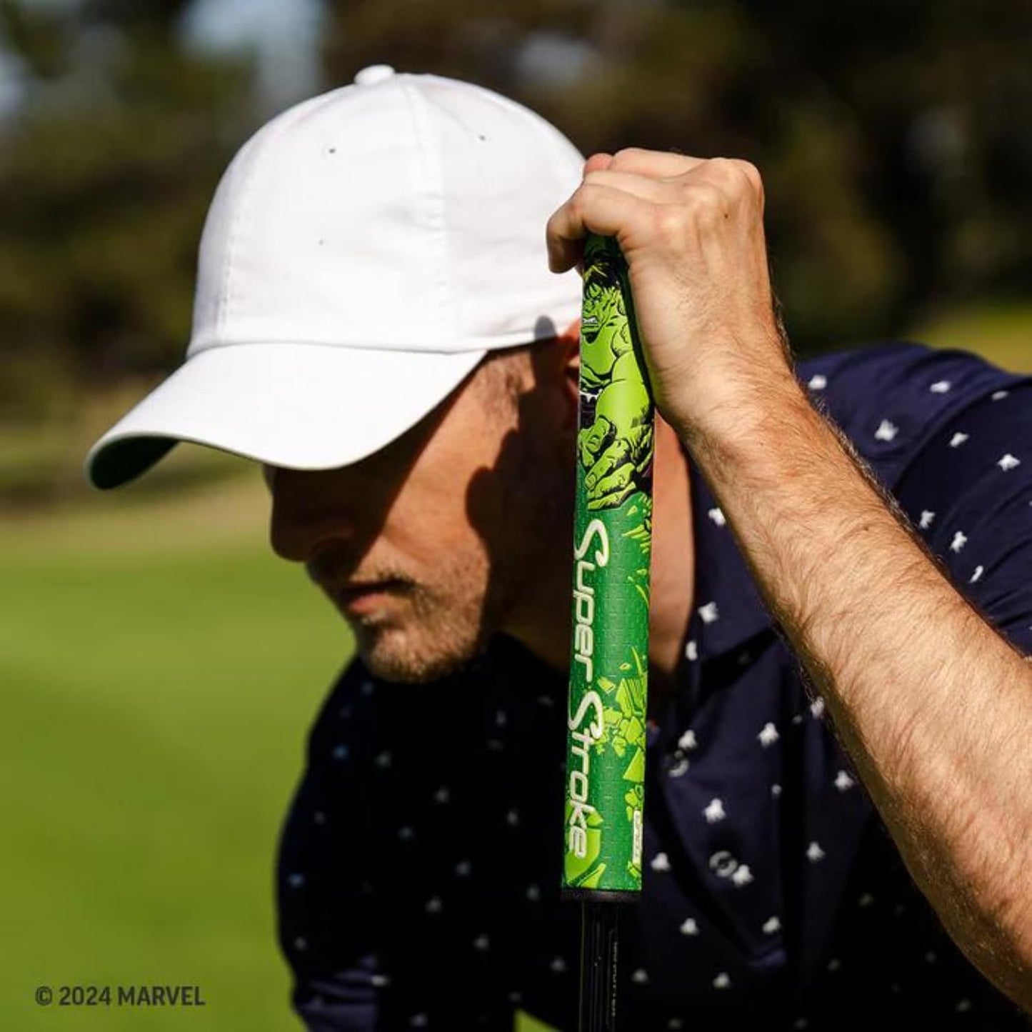Man holding a golf club with a Marvel logo on a blurred golf course background