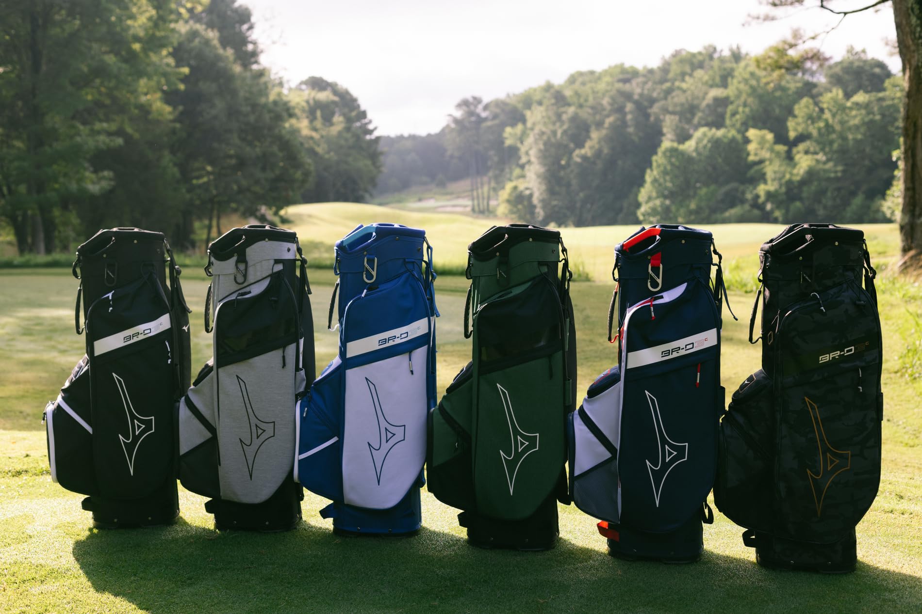 Six golf bags of different colors lined up on a grassy area with trees in the background